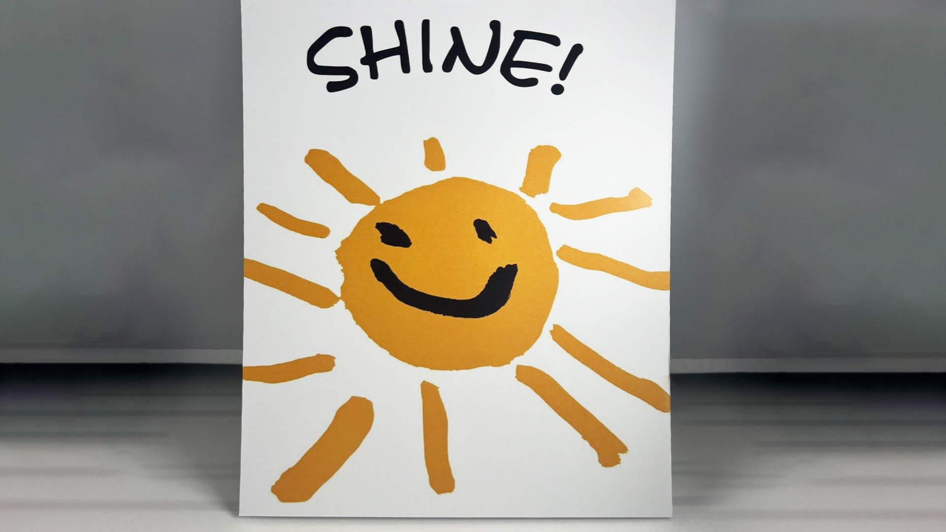 A hand drawn yellow sun with a smiley face, surrounded by sun rays with the word SHINE! written above it.